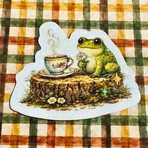 Cute cottagecore frog sticker with holographic sparkle overlay, featuring a green frog holding a daisy beside a teacup on a tree stump, displayed on a warm plaid gingham background.