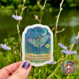 A hand holds a holographic vinyl sticker featuring a whimsical Luna moth beneath a crescent moon, surrounded by lavender, yellow flowers, and twinkling stars. The sticker includes the quote “I dance with the wind and speak with the moon” on a golden scroll. The background shows a lush green field and a calm pond, with soft purple wildflowers in bloom. The person’s nails are painted with a glittery galaxy design, and the Sticker Gypsy Co. logo appears in the bottom right corner.