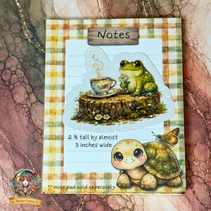 Cute cottagecore frog sticker on a notepad mockup, featuring a frog holding a daisy beside a teacup on a tree stump, with a gingham background and a smiling tortoise illustration, holographic sparkles visible.