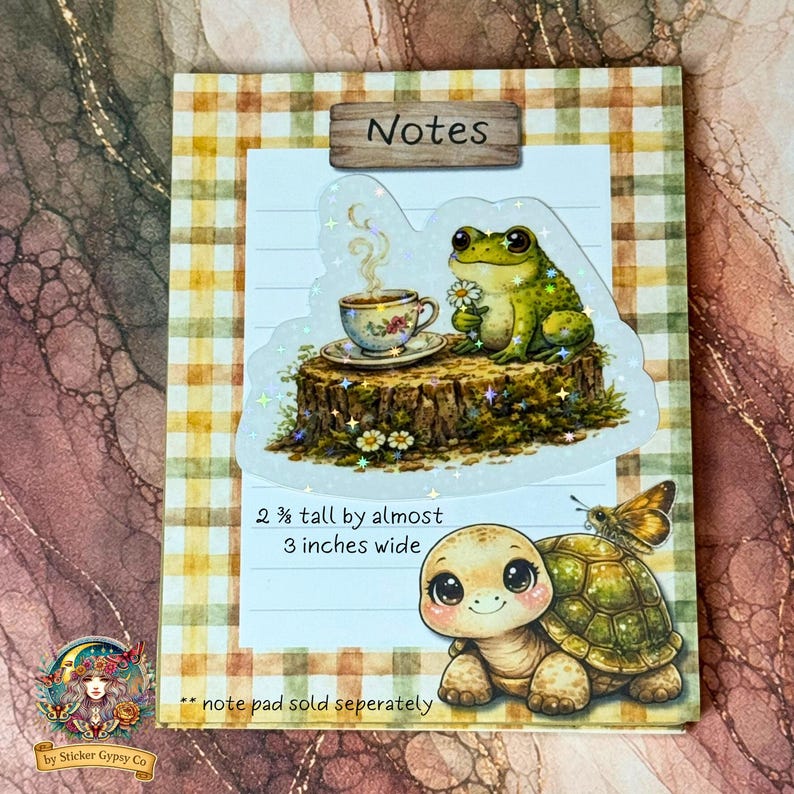 Cute cottagecore frog sticker on a notepad mockup, featuring a frog holding a daisy beside a teacup on a tree stump, with a gingham background and a smiling tortoise illustration, holographic sparkles visible.