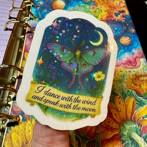 A hand holds a holographic vinyl sticker depicting a luminous Luna moth surrounded by lavender, wildflowers, and a crescent moon in a deep blue starry sky. The sticker sparkles with rainbow holographic stars and includes the quote "I dance with the wind and speak with the moon" on a yellow scroll banner. The background features a colorful galaxy-themed notebook with swirling stars and planets, alongside vibrant