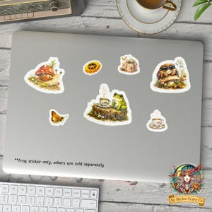Cottagecore frog sticker displayed on a laptop alongside other woodland animal stickers including snail, owl, butterfly, and teacup designs, cozy desk scene with coffee and keyboard.