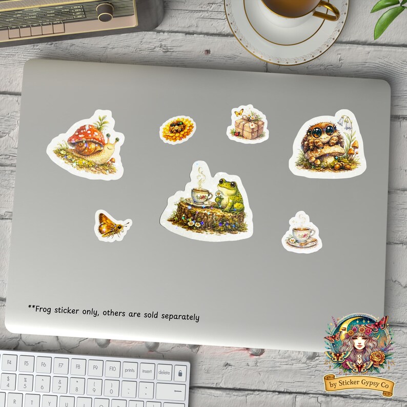 Cottagecore frog sticker displayed on a laptop alongside other woodland animal stickers including snail, owl, butterfly, and teacup designs, cozy desk scene with coffee and keyboard.