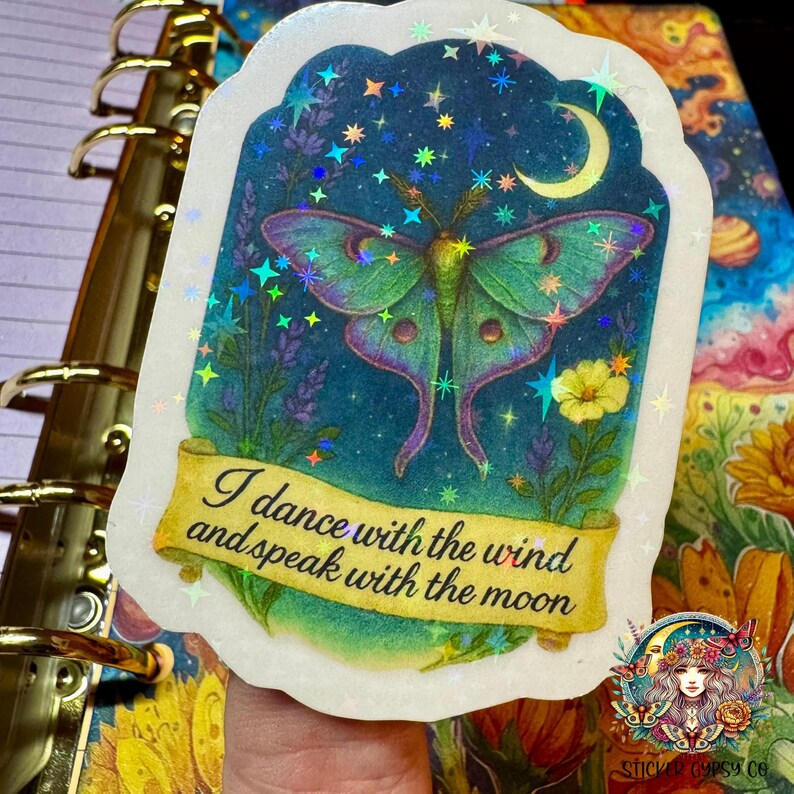 A hand holds a holographic vinyl sticker featuring a glowing green Luna moth under a crescent moon with the phrase "I dance with the wind and speak with the moon" written on a scroll. The background includes purple lavender, yellow wildflowers, and a starry night sky filled with holographic rainbow stars. The sticker rests atop a vibrant cosmic planner and sunflower-themed journal. Sticker Gypsy Co. logo is visible in the bottom right corner.