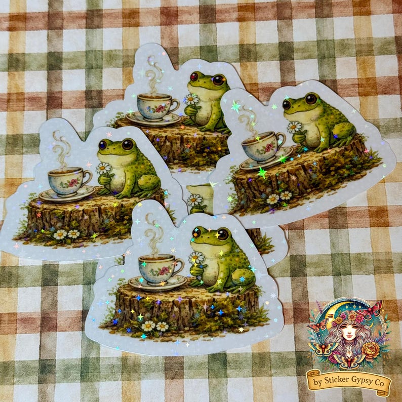 a pile of holographic frog stickers with cottagecore woodland design, showing multiple copies of a frog with teacup and flowers arranged on a neutral gingham background.