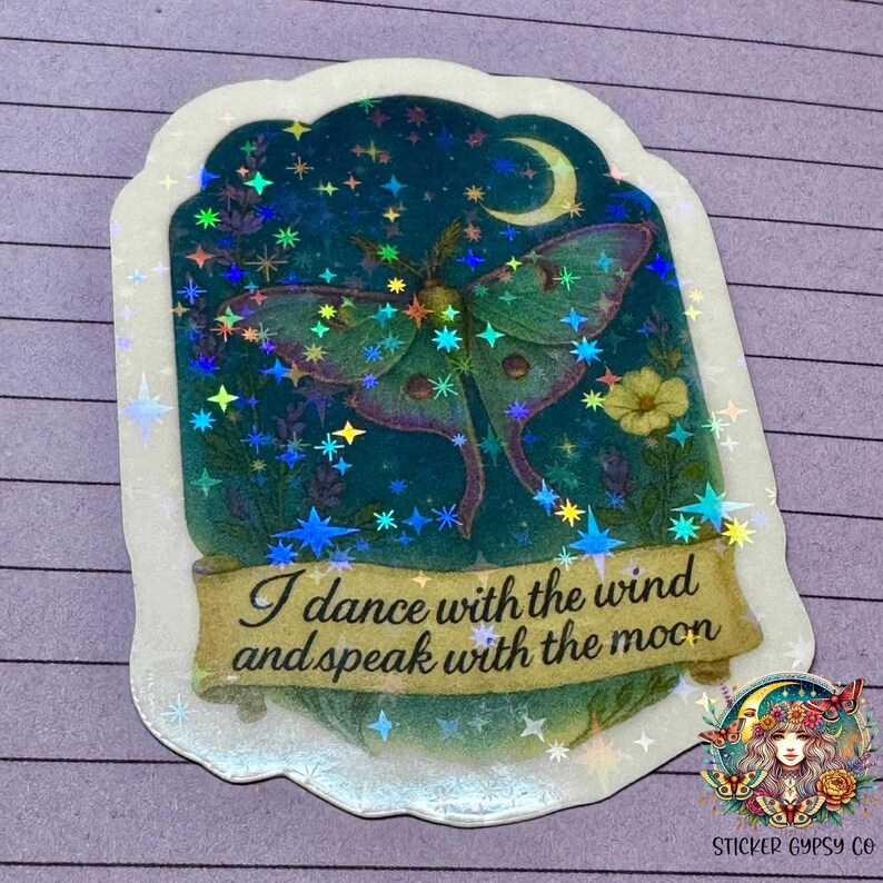 A holographic vinyl sticker featuring a dreamy Luna moth under a crescent moon is shown on a purple lined surface. The design includes lavender, yellow flowers, and a sparkling starry sky, with rainbow holographic stars scattered across the surface. A golden scroll banner at the bottom reads, "I dance with the wind and speak with the moon." The Sticker Gypsy Co. logo is placed in the bottom right corner.