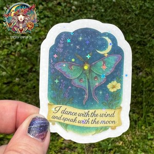 A hand holds a holographic vinyl sticker featuring a vivid green Luna moth with feathered antennae flying beneath a crescent moon. The moth is surrounded by lavender, yellow wildflowers, and a night sky filled with shimmering rainbow holographic stars. The quote “I dance with the wind and speak with the moon” is written on a golden scroll at the bottom. The background is grassy and outdoors, and the Sticker Gypsy Co. logo appears in the upper left corner.