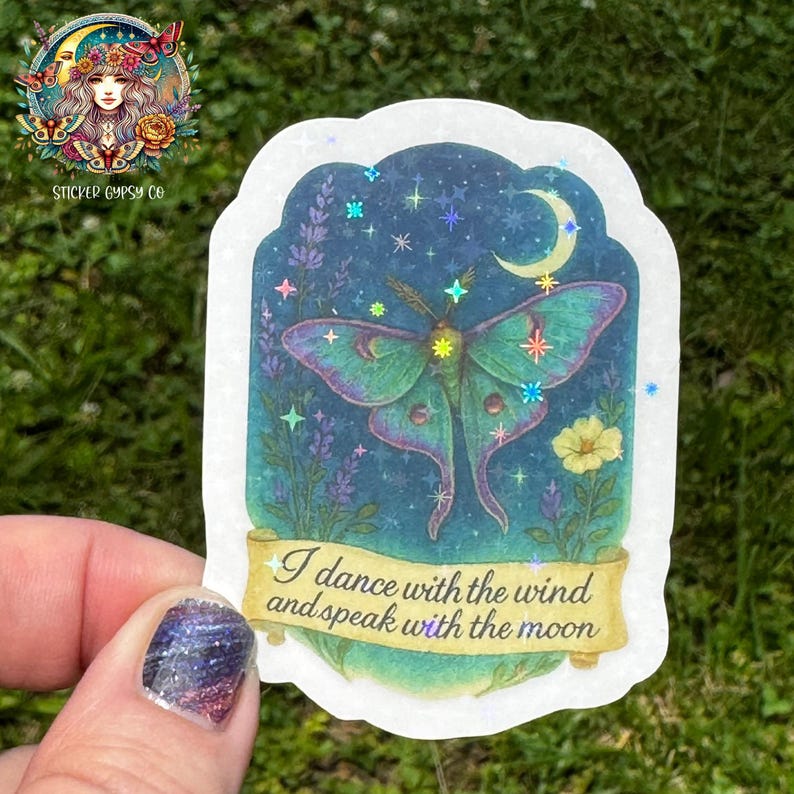 A hand holds a holographic vinyl sticker featuring a vivid green Luna moth with feathered antennae flying beneath a crescent moon. The moth is surrounded by lavender, yellow wildflowers, and a night sky filled with shimmering rainbow holographic stars. The quote “I dance with the wind and speak with the moon” is written on a golden scroll at the bottom. The background is grassy and outdoors, and the Sticker Gypsy Co. logo appears in the upper left corner.