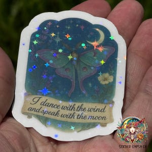 A hand holds a holographic vinyl sticker featuring a magical Luna moth with feathered antennae beneath a crescent moon, surrounded by lavender and yellow wildflowers against a starry night sky. Sparkling holographic rainbow stars shimmer across the design. A golden scroll at the bottom reads, “I dance with the wind and speak with the moon.” The Sticker Gypsy Co. logo is placed in the lower right corner.