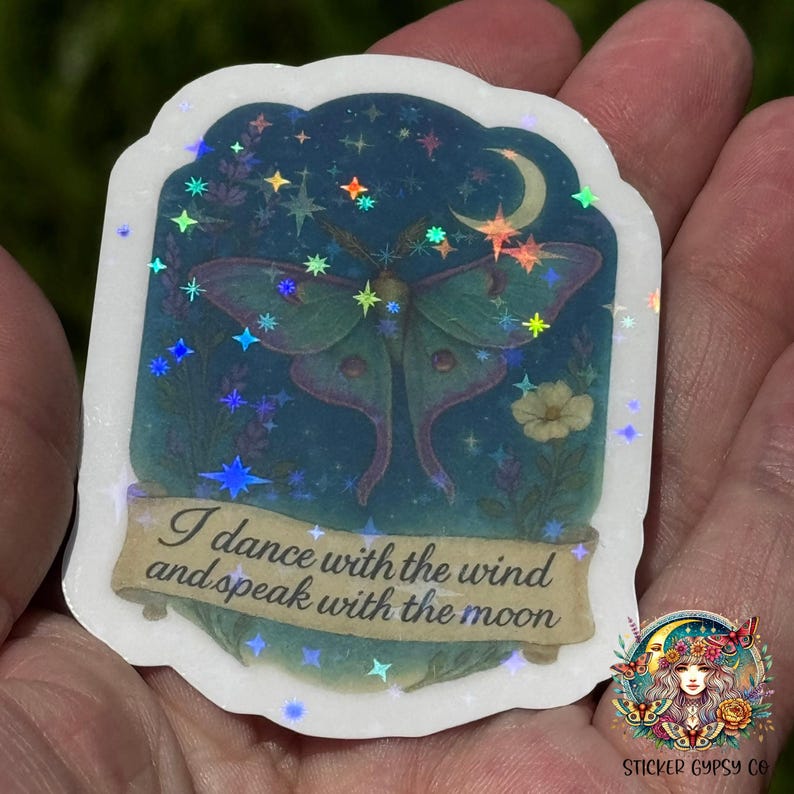 A hand holds a holographic vinyl sticker featuring a magical Luna moth with feathered antennae beneath a crescent moon, surrounded by lavender and yellow wildflowers against a starry night sky. Sparkling holographic rainbow stars shimmer across the design. A golden scroll at the bottom reads, “I dance with the wind and speak with the moon.” The Sticker Gypsy Co. logo is placed in the lower right corner.