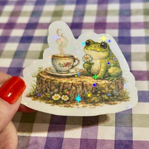 Detailed close-up of holographic frog sticker with rainbow sparkle effect, showing a green frog holding a flower next to a steaming teacup on a rustic stump.