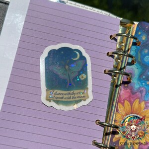 A holographic vinyl sticker featuring a mystical Luna moth under a crescent moon is placed on a lavender-lined journal page inside a binder. The sticker includes lavender flowers, yellow blooms, and starry sky details with holographic rainbow stars. The quote “I dance with the wind and speak with the moon” appears on a golden scroll at the bottom. The edge of the journal shows a vibrant cosmic floral design with planets and sunflowers. The Sticker Gypsy Co. logo is displayed in the right corner.