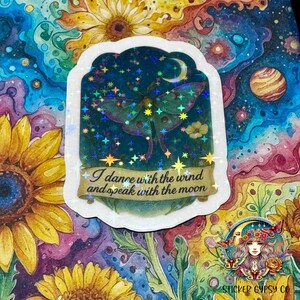 A holographic vinyl sticker featuring a glowing Luna moth beneath a crescent moon is placed on a vibrant cosmic floral background. The design includes lavender, yellow flowers, and a starry night sky, accented by shimmering rainbow holographic stars. The quote “I dance with the wind and speak with the moon” is written on a golden scroll banner at the bottom. Surrounding the sticker are vivid watercolor-style sunflowers and swirling galaxies.