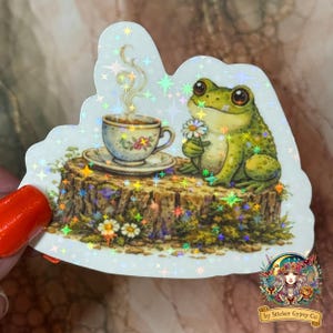K&ouml;nnte beinhalten: Ein skurriler Aufkleber mit einem gr&uuml;nen Frosch, der eine G&auml;nsebl&uuml;mchen h&auml;lt, auf einem Baumstumpf neben einer Teetasse sitzt. Die Tasse und Untertasse haben florale Designs. Der Aufkleber hat ein holografisches Finish mit sternf&ouml;rmigen Akzenten.