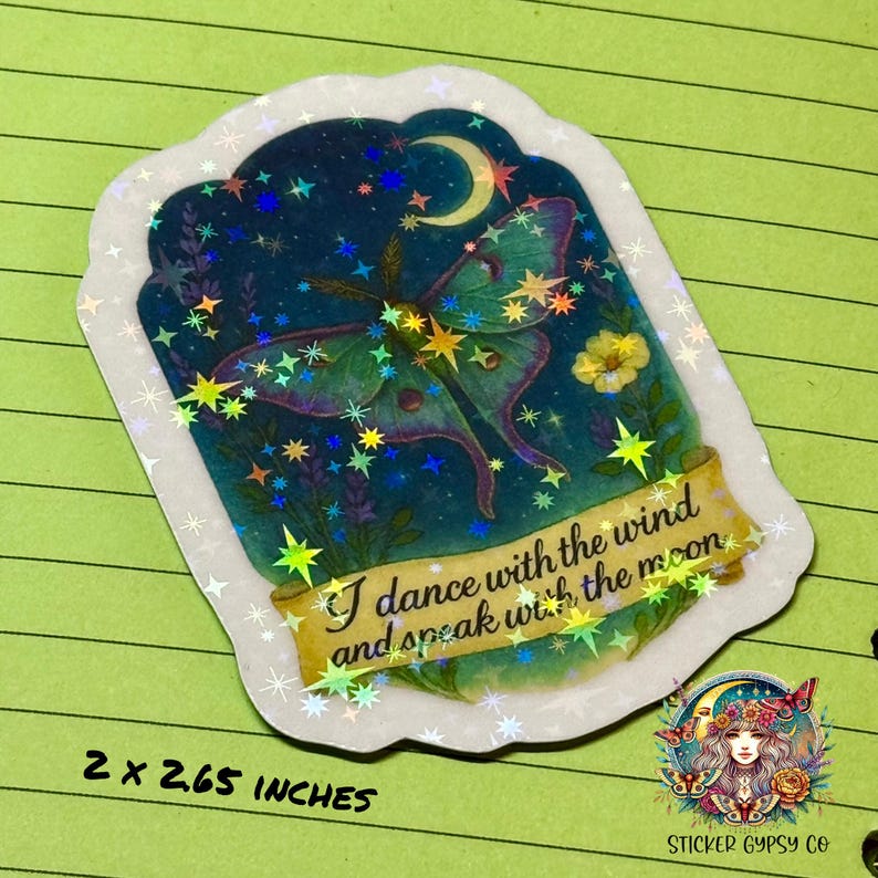 A 2 x 2.65 inch holographic vinyl sticker is shown on a bright green lined background. The sticker features a glowing green Luna moth beneath a crescent moon, surrounded by lavender and yellow flowers against a starry night sky. The text reads, "I dance with the wind and speak with the moon" on a golden scroll banner. Rainbow holographic stars shimmer across the design. The Sticker Gypsy Co. logo appears in the bottom right corner.