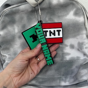 May include: A green and black Creeper and red and white TNT luggage tag with the text "YOUR NAME". The tag is attached to a gray tie-dye backpack with a silver wire.
