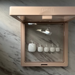 Puede incluir: Un juego de seis uñas postizas cuadradas blancas en un estuche de plástico transparente con tapa rosa. Las uñas están ordenadas por tamaño, con la uña más pequeña a la izquierda y la más grande a la derecha.