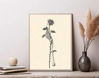 May include: A framed wall art print featuring a black and white ink illustration of a skeletal hand holding a single rose with a thorny stem. The artwork is centered on a light cream background, showcasing detailed bone structure and floral petals.