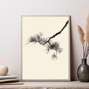 May include: A rectangular framed wall art print featuring a black ink illustration of a pine tree branch with needles on a solid cream-colored background. The branch extends diagonally from the upper right corner toward the center of the frame.