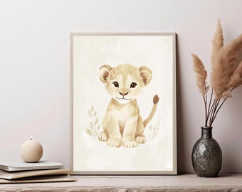 May include: A framed wall art print featuring a watercolor illustration of a light tan lion cub sitting upright. The cub has large dark eyes, a small nose, and a simple tail. The background is a soft cream color with subtle plant outlines.