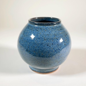 May include: A round, blue ceramic vase with a speckled texture. The vase has a wide, rounded body and a narrow opening. The base is a lighter shade of blue. The vase is approximately 7 inches tall and 8 inches wide.