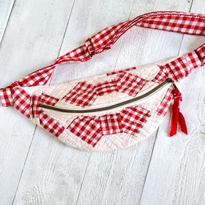May include: A quilted fanny pack featuring a red and white gingham pattern on a light pink background. The bag has an adjustable strap in the same gingham fabric, a metal zipper closure, and a red fabric tassel attached to the zipper pull.