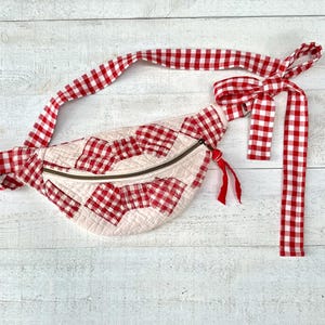 May include: A quilted fanny pack featuring a red and white gingham check pattern. The bag has a curved shape with a metal zipper closure, a matching long fabric strap that ties into a bow, and a small red fabric pull attached to the zipper.