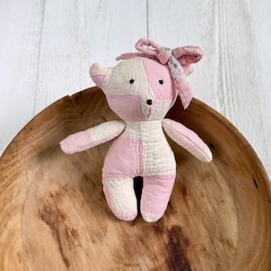 May include: A small, handmade stuffed bear toy crafted from quilted fabric patches in alternating shades of soft pink and cream. The bear features simple black stitched eyes and nose, with a decorative floral patterned fabric bow tied on its head.