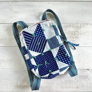 May include: A quilted backpack with a patchwork design in white and navy blue. The backpack features a variety of geometric patterns and is accented with teal green ties. The straps are teal green with a white pattern.