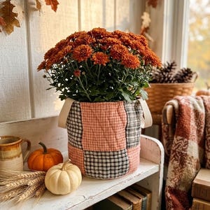 May include: A round, quilted fabric planter with a patchwork pattern of orange and black gingham squares, featuring two cream-colored fabric handles. The planter holds a dense, bushy plant with numerous small, vibrant orange chrysanthemum flowers.