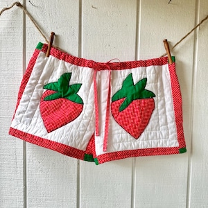 May include: White quilted shorts with red polka dot trim and two large strawberry appliqués. The strawberries are red with white polka dots and green leaves. A red ribbon drawstring is visible.