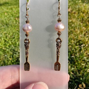 Könnte beinhalten: Ohrringe zum Hängen mit einer hellrosa Perle und einem bronzefarbenen Gabelanhänger. Die Ohrringe haben goldfarbene Ohrhaken und kleine Perlen. Sie werden auf einem transparenten, mattierten Acrylständer vor einem verschwommenen grünen Hintergrund präsentiert.