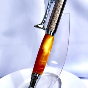 May include: A handcrafted pen with a chrome clip and accents. The pen's barrel is a vibrant blend of orange and yellow, with a detailed design on the upper portion. The pen is displayed on a clear acrylic stand.