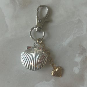 May include: A silver-colored keychain with a seashell charm and a smaller gold-colored seashell charm. The keychain has a lobster clasp for easy attachment to bags or keys. The charms are detailed and add a coastal touch.