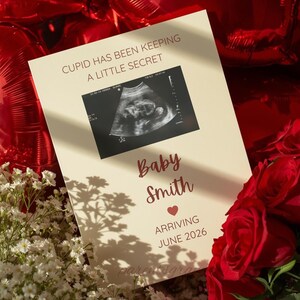 Peut inclure: Une carte d'annonce crème avec le texte "Cupidon Garde un Petit Secret" et "Baby Smith" en bordeaux. Une image d'échographie est présentée, avec "Arrivée Juin 2026" en dessous. Des roses rouges et des fleurs blanches entourent la carte.