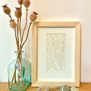 May include: A framed white paper cut art piece with a natural wood frame. The art piece features a detailed design of a tree branch with intricate lines and curves. The frame is sitting on a wooden surface with a vase of dried poppy pods, a piece of driftwood, and a few stones.
