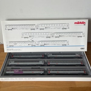 May include: A Märklin HO scale model train set with six cars. The cars are gray with red accents and feature the words "Railjet" and "ÖBB" on the side. The set is in its original packaging, which is white with blue and red accents. The packaging also includes a diagram of the train set.