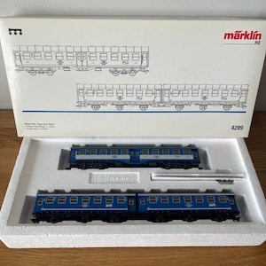 Puede incluir: Un set de tren modelo Märklin a escala HO con dos coches de pasajeros azules. Los coches tienen molduras blancas y el texto "Express-Bahn" en el lateral. El set está en su embalaje original e incluye una caja con un diagrama del set de tren.