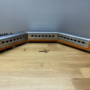 May include: A vintage toy train set with three cars, including an engine car, a passenger car, and a caboose. The train is made of metal and painted in orange and silver. The train is on a wooden surface.