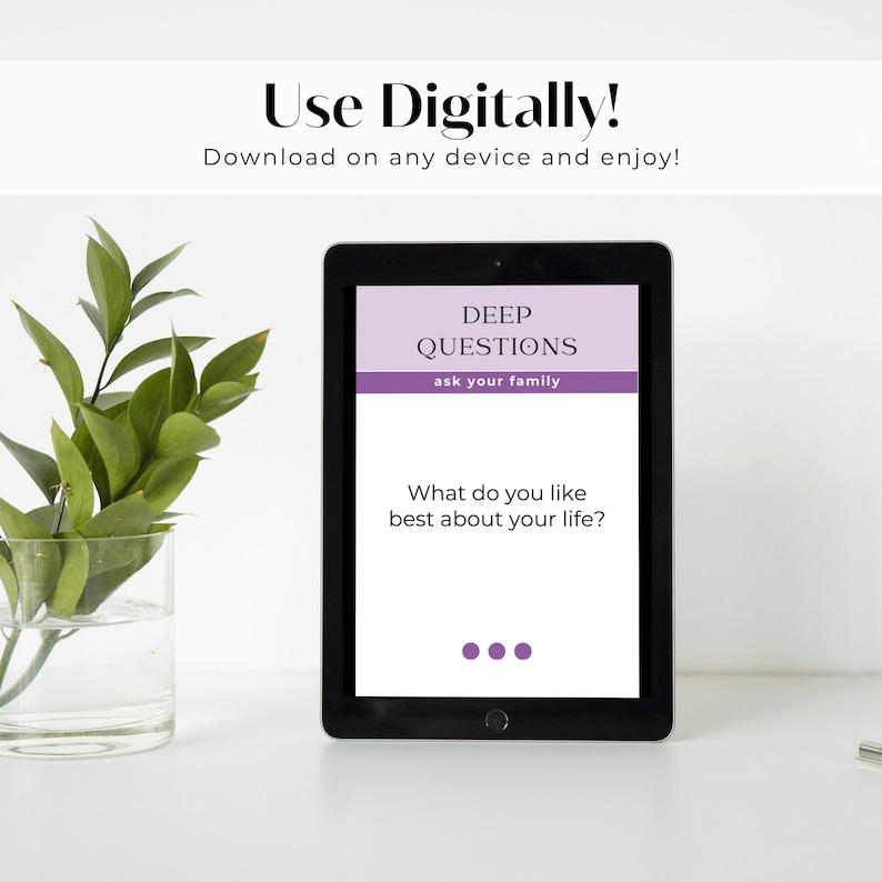 May include: A black tablet with a white screen displaying a digital card with the text "DEEP QUESTIONS ask your family" and the question "What do you like best about your life?" The card has a purple background and white text. The tablet is sitting on a white surface next to a clear glass vase with a green plant.