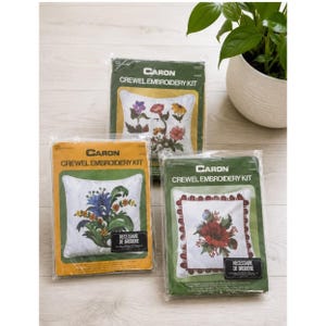 May include: Three Caron Crewel Embroidery Kits in plastic packaging. Each kit features a white square fabric panel with floral designs, including blue, red, and multi-colored flowers. The packaging displays the brand name and text.