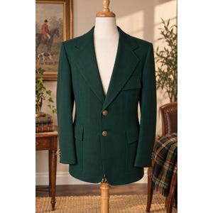 May include: A dark forest green blazer featuring a notched lapel, two gold-toned buttons at the center front, and two flap pockets at the waist. The jacket includes a single chest pocket with a flap and long sleeves with gold-toned buttons.