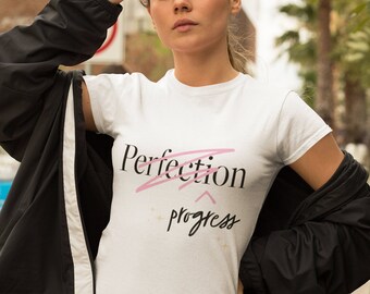 May include: White t-shirt with the words "Perfection" crossed out and replaced with "progress" in a stylish font. The shirt is worn with a black jacket and sunglasses.