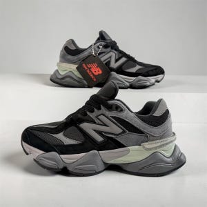 Op de afbeelding: Een paar zwart-grijze New Balance sneakers met mesh en suède panelen, zwarte veters en een dikke grijze tussenzool met lichtgroene accenten. Eén sneaker is voorzien van een zwart New Balance merklabel aan de veteroogjes.