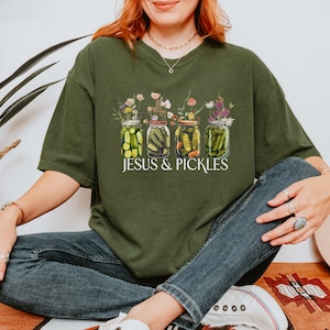 Könnte beinhalten: Dunkelgrünes T-Shirt mit einer Grafik von vier Gläsern, die mit Essiggurken und Blumen gefüllt sind. Der Text "JESUS & PICKLES" ist darunter in Weiß gedruckt. Das Shirt ist ein bequemer, lässiger Stil.