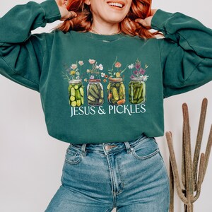 Könnte beinhalten: Dunkelgrünes Sweatshirt mit einer Grafik von vier Gläsern mit eingelegten Gurken, die mit Blumen bedeckt sind. Der weiße Text "JESUS & PICKLES" ist darunter aufgedruckt. Das Sweatshirt ist einfarbig.