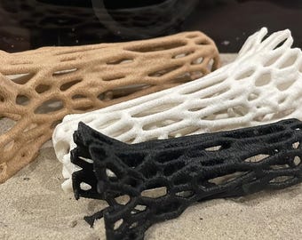 May include: Three decorative, hollow, branch-like structures in tan, white, and black. The objects have an open, web-like design and are resting on a bed of sand. The objects appear to be 3D printed.