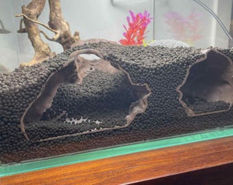 May include: A glass aquarium with a brown wooden base. The tank contains dark gravel, driftwood, and decorative cave structures. Artificial plants in pink and red add color to the aquatic environment.