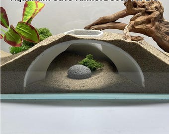 May include: Aquarium Cave Tunnel Décor TYPE C. A white, arched tunnel structure is partially buried in sand, creating a cave-like environment. A small, gray stone and green plant life are inside the tunnel. The text "Aquarium Cave Tunnel Décor TYPE C" is at the top.