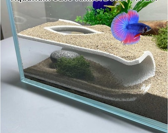 May include: An aquarium with a white cave tunnel decor and a colorful betta fish. The tank features a sand substrate, a rock with green moss, and artificial plants. Text on the image reads "Aquarium Cave Tunnel Décor TYPE A."